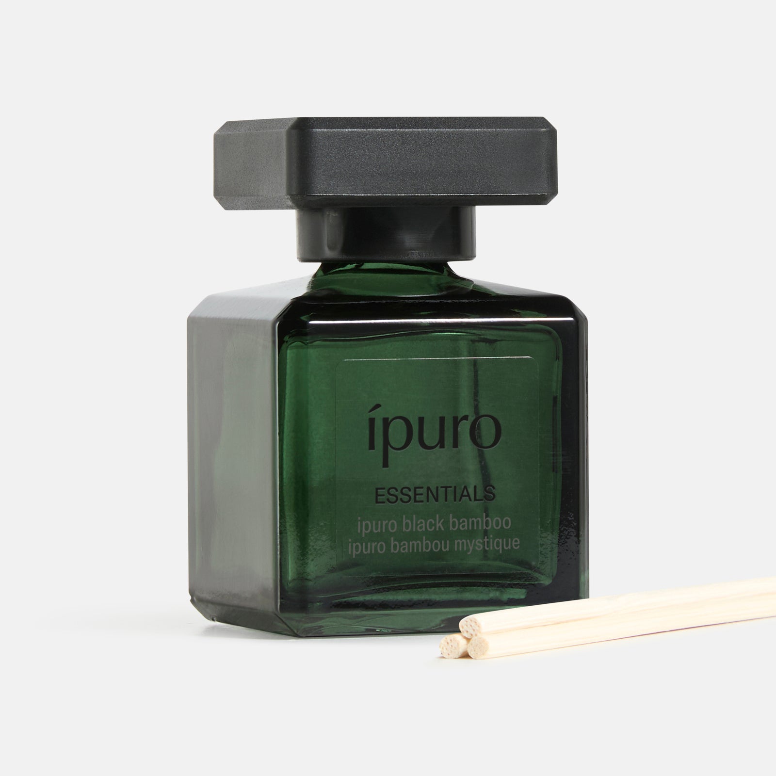 Ein quadratischer, dunkelgrüner Glasflakon von ipuro Raumduft black bamboo mit schwarzer Kappe steht neben zwei hölzernen Diffusorstäben, die frische grüne und holzige Noten auf einem hellen Hintergrund hervorrufen. Marke: ipuro.