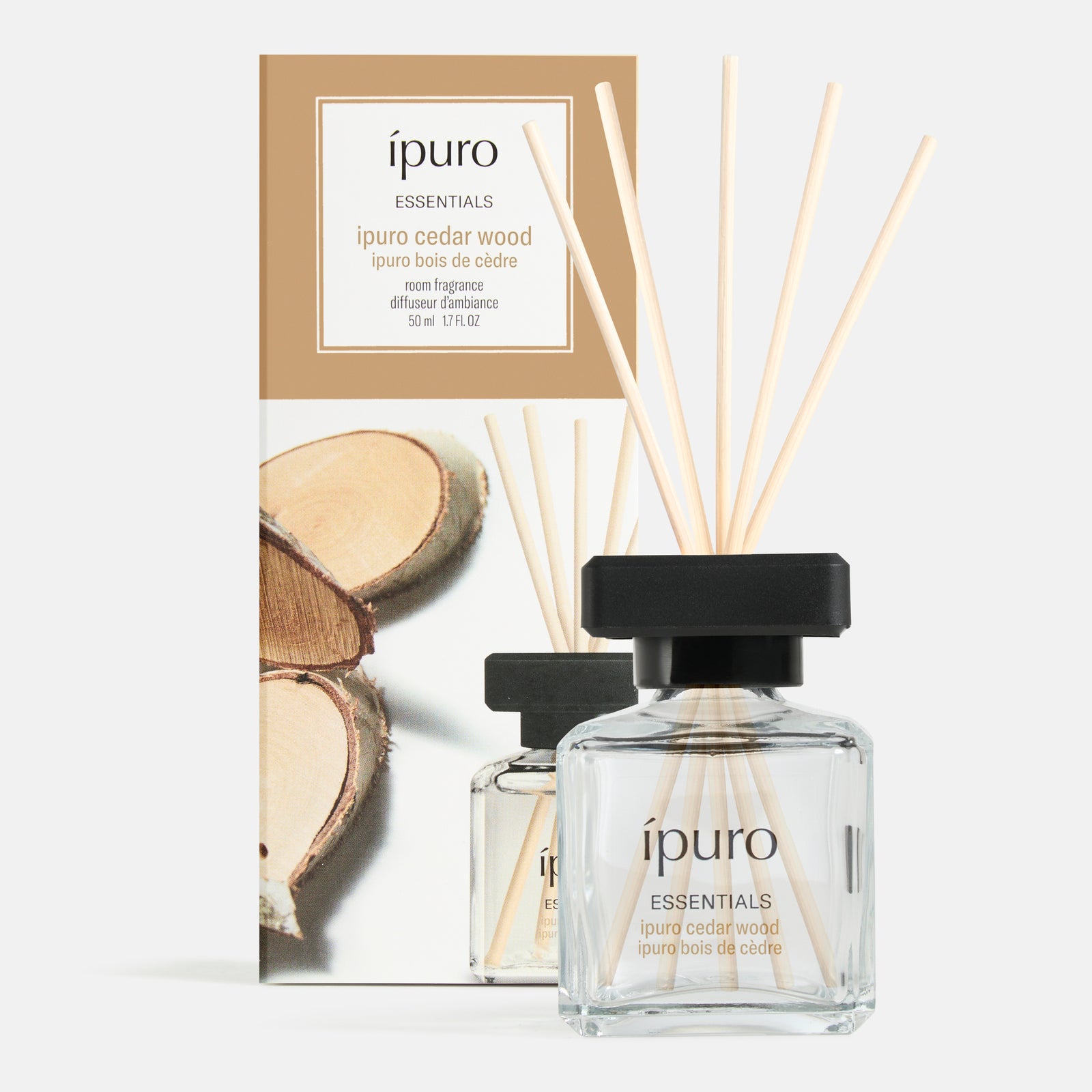 Ein klarer Glasflakon mit der Aufschrift ipuro Raumduft Zedernholz mit schwarzer Kappe und Schilfrohrstäbchen steht vor seiner Verpackung, die Holzscheiben zeigt und den holzigen Duft mit subtilen fruchtigen Noten von ipuro widerspiegelt.