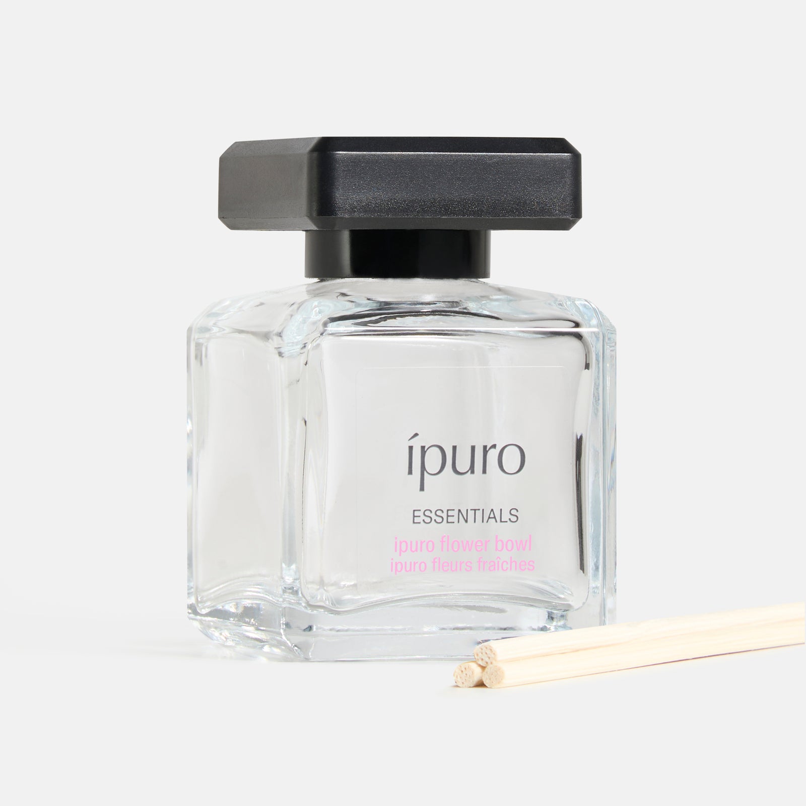 Ein klarer Glasflakon von ipuro Raumduft lovely flowers by ipuro mit einer schwarzen quadratischen Kappe und zwei leichten Holzstäbchen für den Diffusor wird vor einem schlichten weißen Hintergrund präsentiert.