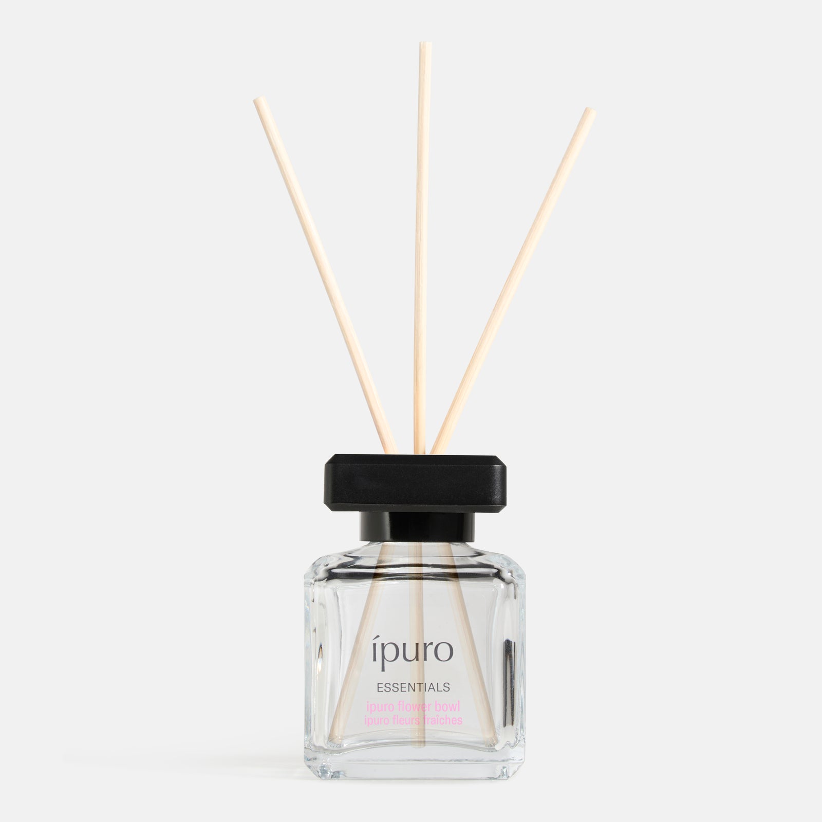 Eine klare Glasflasche von ipuro Raumduft lovely flowers Diffusor mit einer schwarzen Kappe und drei hölzernen Schilfrohr, gefüllt mit duftenden Flüssigkeit von der Marke ipuro, vor einem schlichten weißen Hintergrund gesetzt.