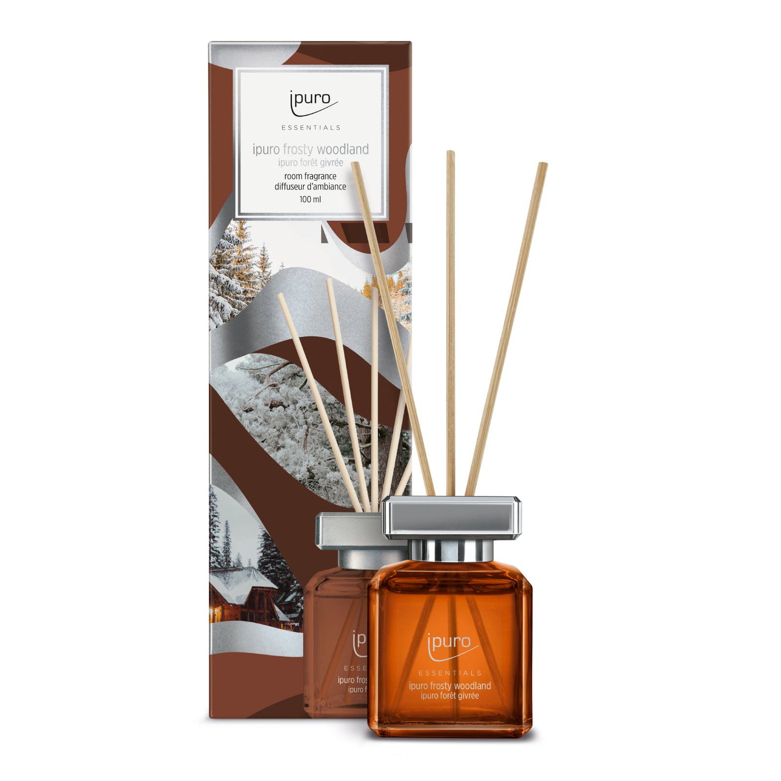 Ein brauner Glasdiffusor mit Schilfrohrstäbchen, beschriftet mit ipuro Essentials frosty woodland, steht vor einer Verpackung mit verschneiten Wäldern und Hütten. Das Set von ipuro enthält 100 ml des Raumdufts.