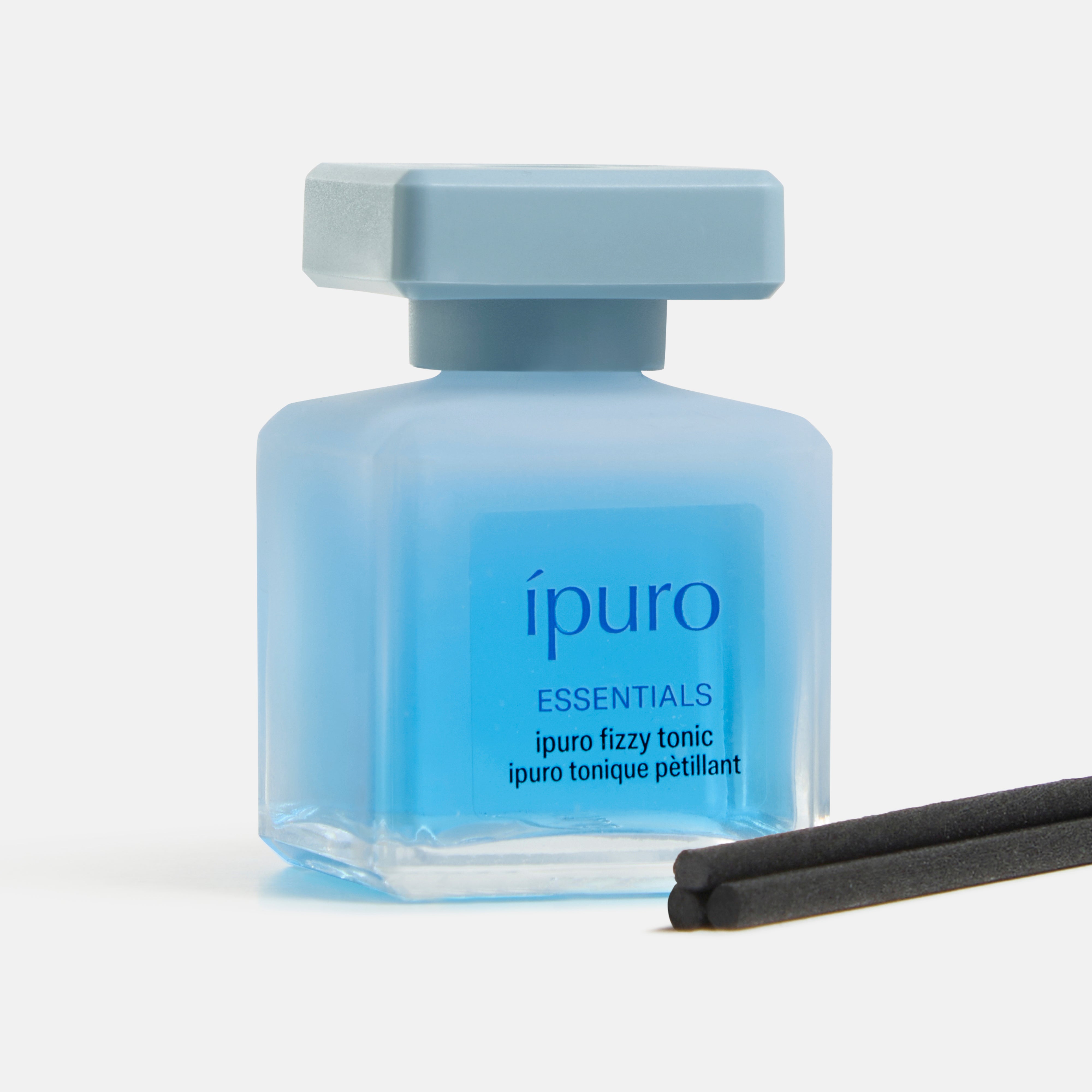 Eine quadratische, hellblaue Flasche ESSENTIALS ipuro fizzy tonic von ipuro, gefüllt mit blauer Flüssigkeit und zwei schwarzen Schilfrohrstäbchen davor, erinnert an einen erfrischenden Gin-Duft mit Wacholder-Noten, der sich vor einem weißen Hintergrund abhebt.