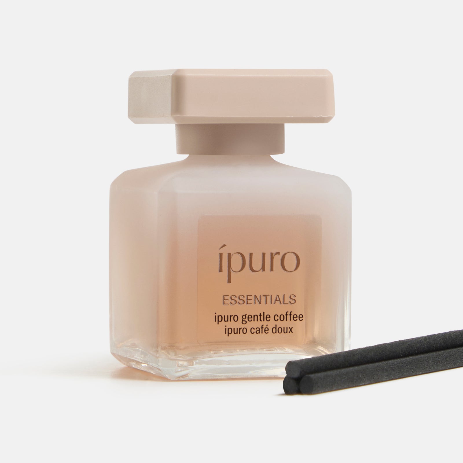 Ein Milchglasflakon von ESSENTIALS ipuro gentle coffee von ipuro mit beigem Verschluss und Etikett wird mit zwei schwarzen Diffusorstäbchen gezeigt. Die Szene erinnert an subtile Mandel- und Vanille-Noten vor einem weißen Hintergrund.
