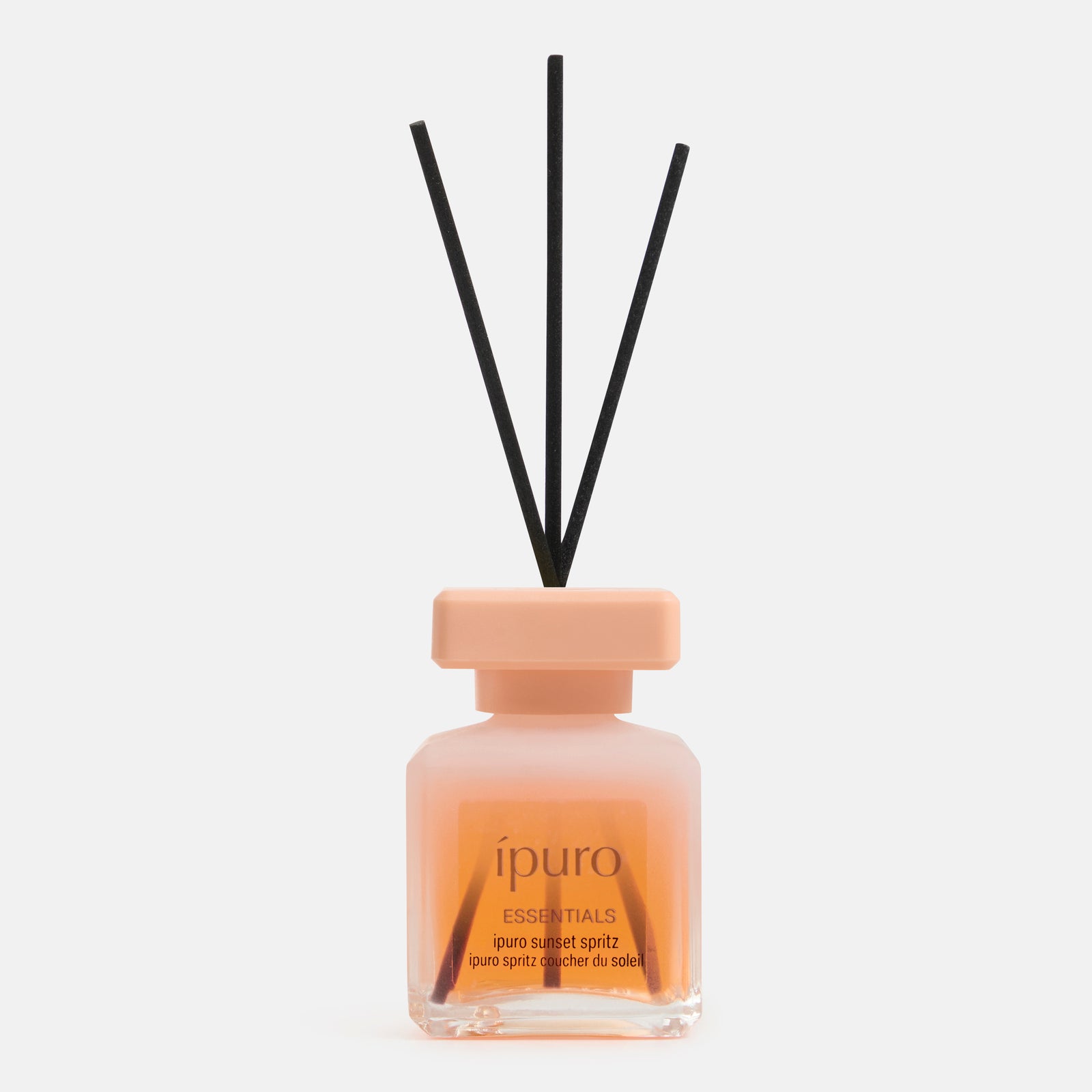 Ein klarer Glasdiffusor mit der Aufschrift "ipuro ESSENTIALS sunset spritz" mit orangefarbener Flüssigkeit und drei schwarzen Schilfrohren von ipuro erinnert an einen lebendigen Sommerabend-Duft wie ein erfrischender Aperitif, der auf einem schlichten weißen Hintergrund präsentiert wird.