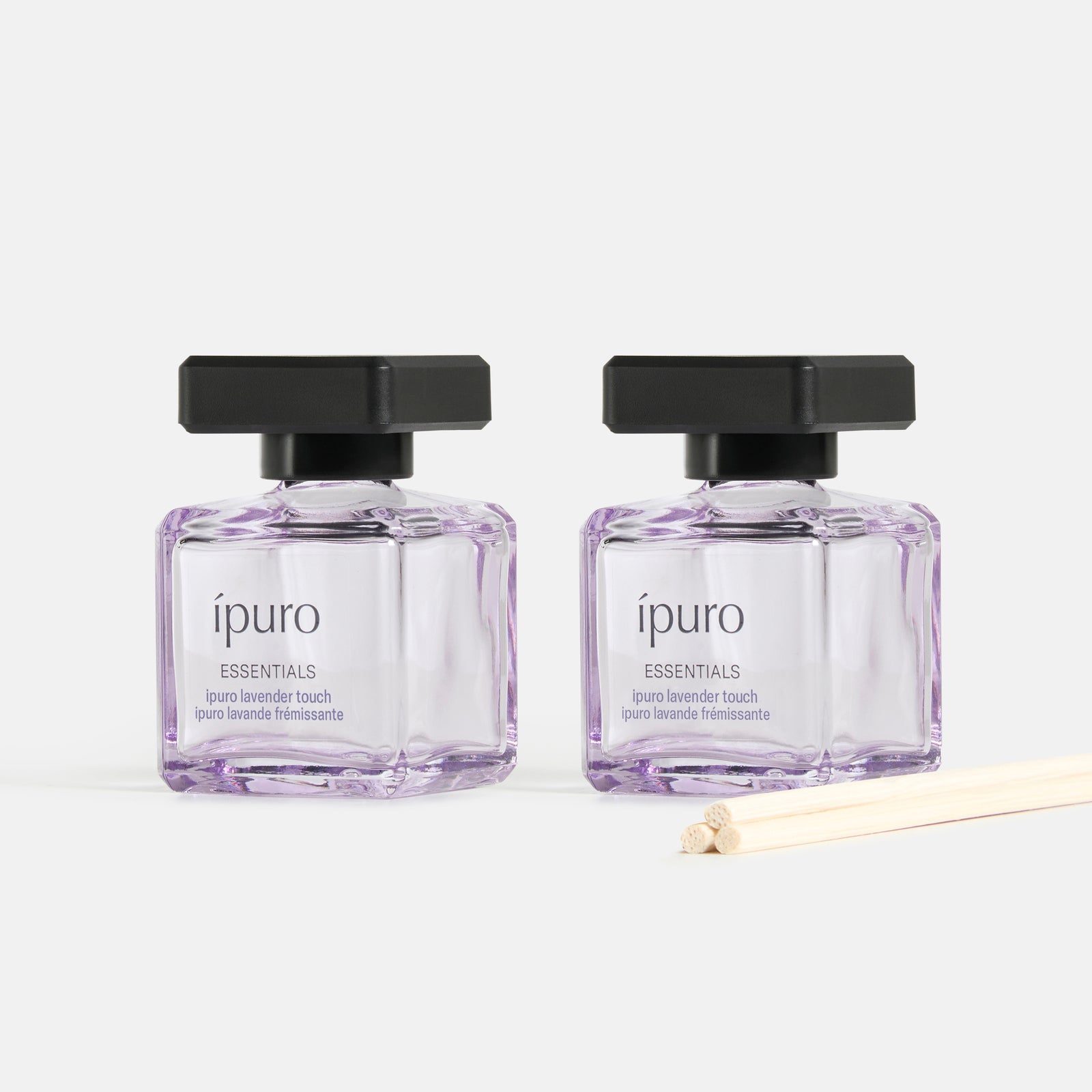Zwei quadratische Glasflaschen mit schwarzem Deckel, gefüllt mit hellvioletter Flüssigkeit, beschriftet mit ipuro Raumduft-Set lavender touch by ipuro, und davor ein Bündel Schilfrohr-Diffusorstäbchen, ausgestellt vor einem weißen Hintergrund.