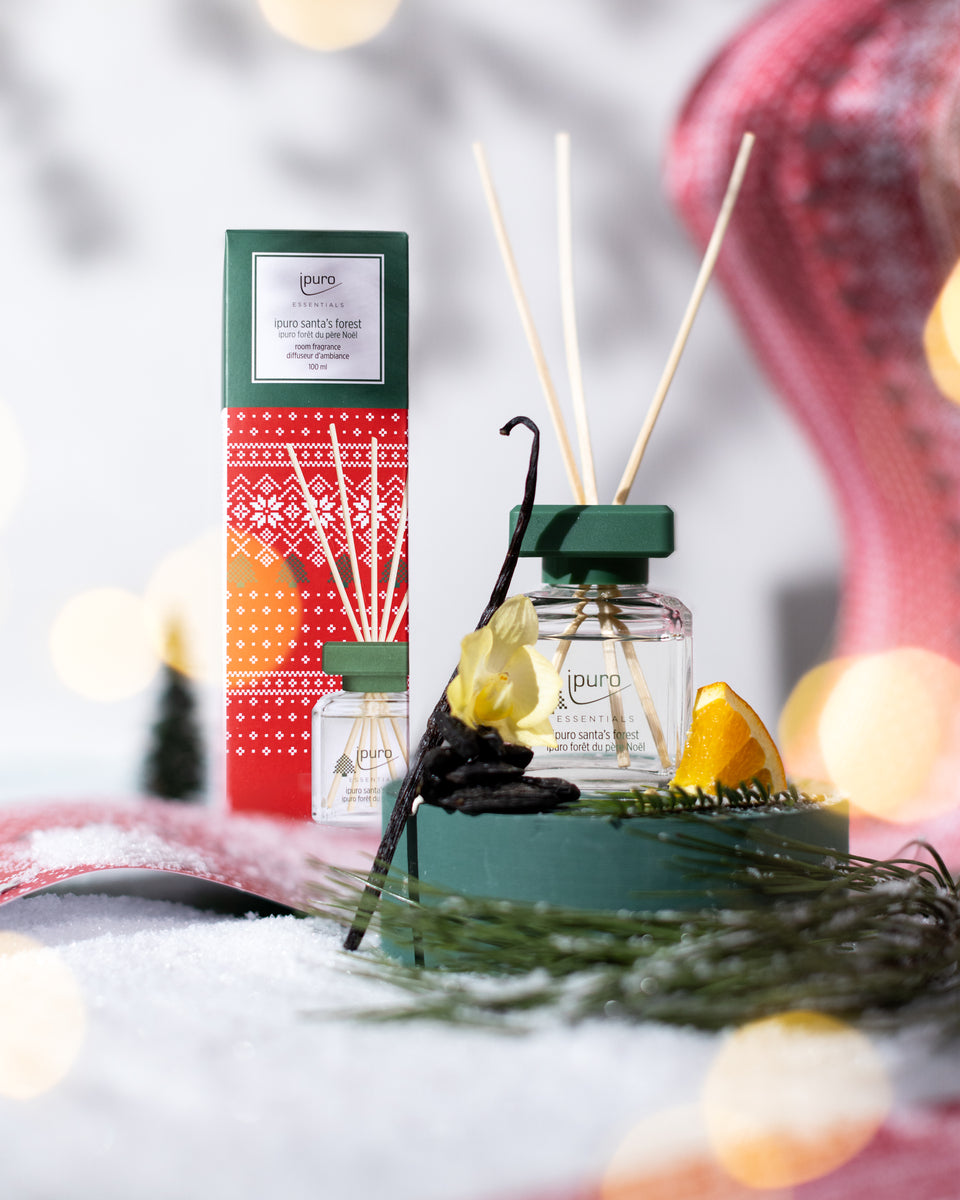 Der ipuro ESSENTIALS Raumduft santa's forest präsentiert sich in einer festlichen grünen und roten Verpackung mit einer dekorativen Vanilleschote, einem Tannenzapfen, einer gelben Blume und einer Orangenscheibe auf Schnee. Genießen Sie gemütliche Weihnachtsduft-Vibes mit seinem charakteristischen Hauch von Fichtenwald.