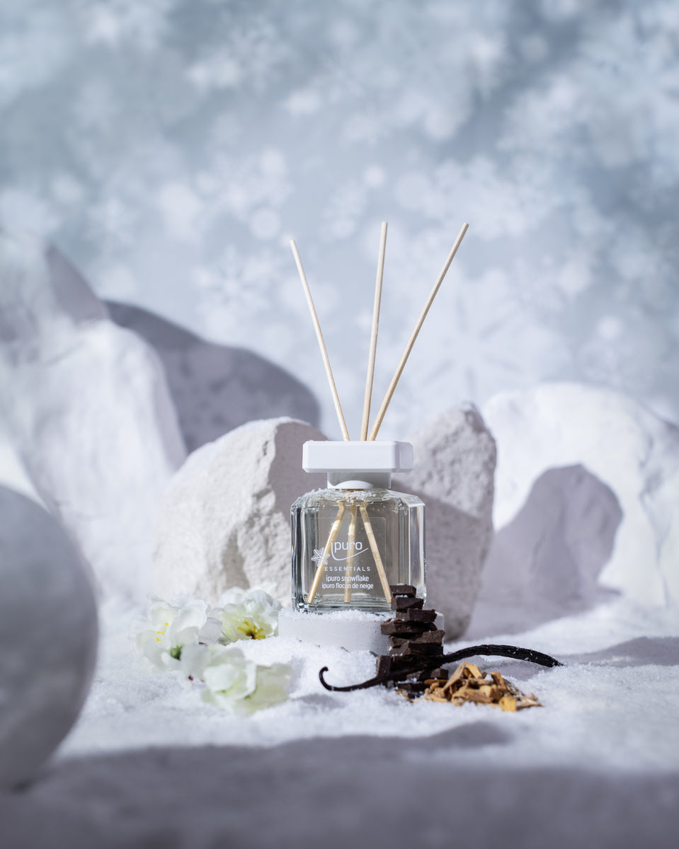 Der ipuro ESSENTIALS Raumduft Snowflake mit weißer Kappe und drei Holzstäbchen ist auf verschneitem Boden zwischen weißen Blumen, Vanilleschoten, Mandelblüten, getrockneten Blütenblättern, weißen Steinen und einem Hintergrund mit Schneeflockenmuster abgebildet.