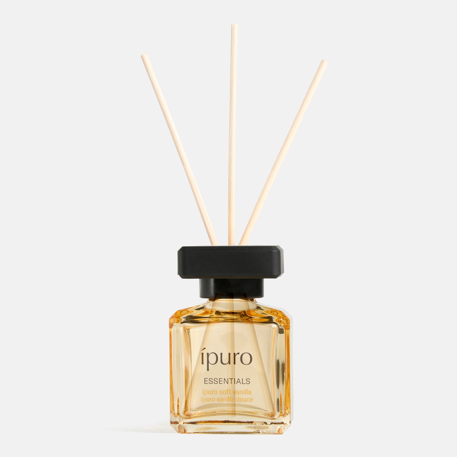 Eine Glasflasche ipuro Raumduft soft vanilla reed diffuser mit hellgelber Flüssigkeit, drei Holzblättern und einer schwarzen Kappe ist vor einem schlichten weißen Hintergrund abgebildet.