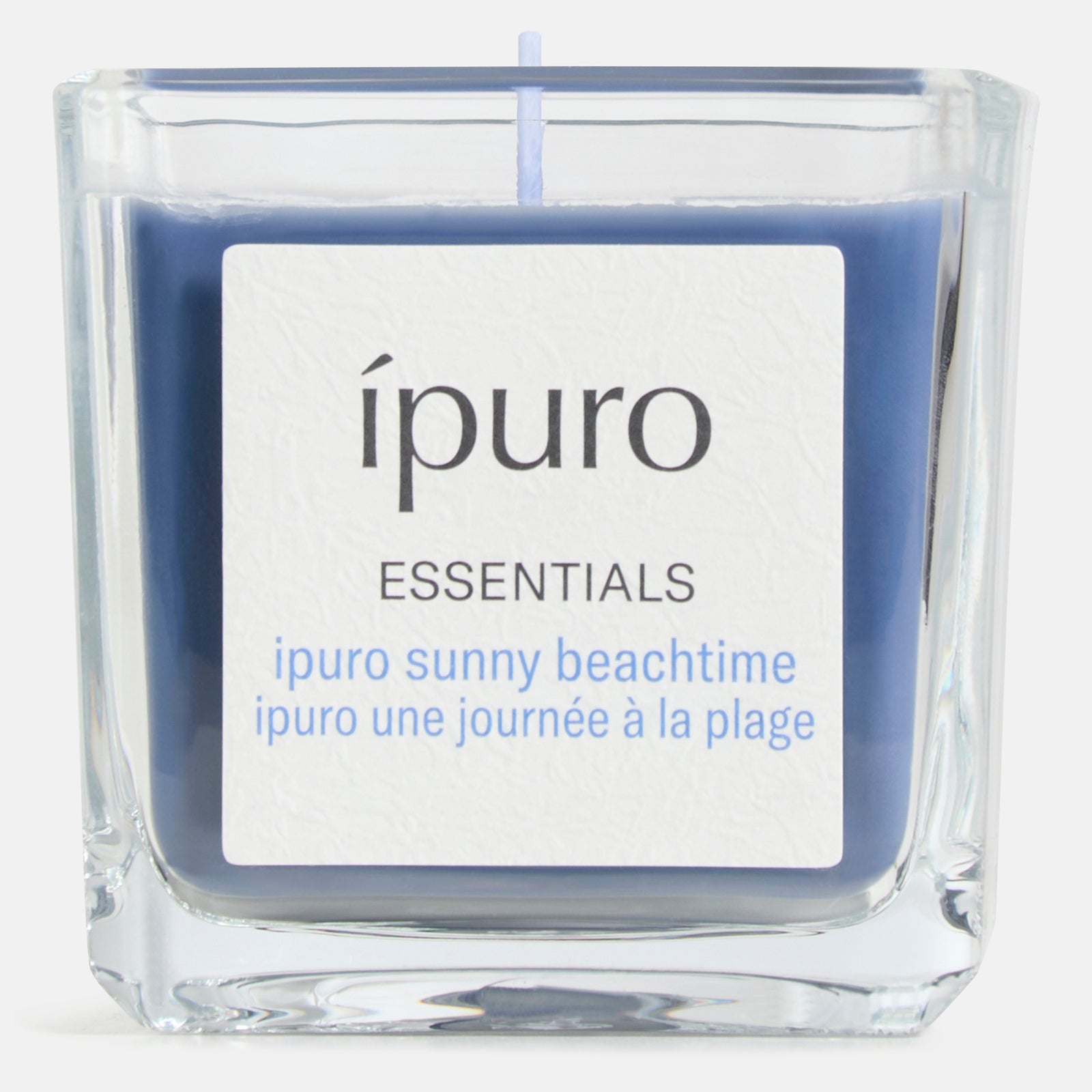 Die ipuro Duftkerze sunny beachtime ist eine blaue Kerze in einem klaren, quadratischen Glas mit den Aufschriften "ipuro ESSENTIALS" und "ipuro sunny beachtime" - perfekt für Sonne, Strand & Sommer - sowie dem französischen "ipuro une journée à la plage" auf einem weißen Etikett.