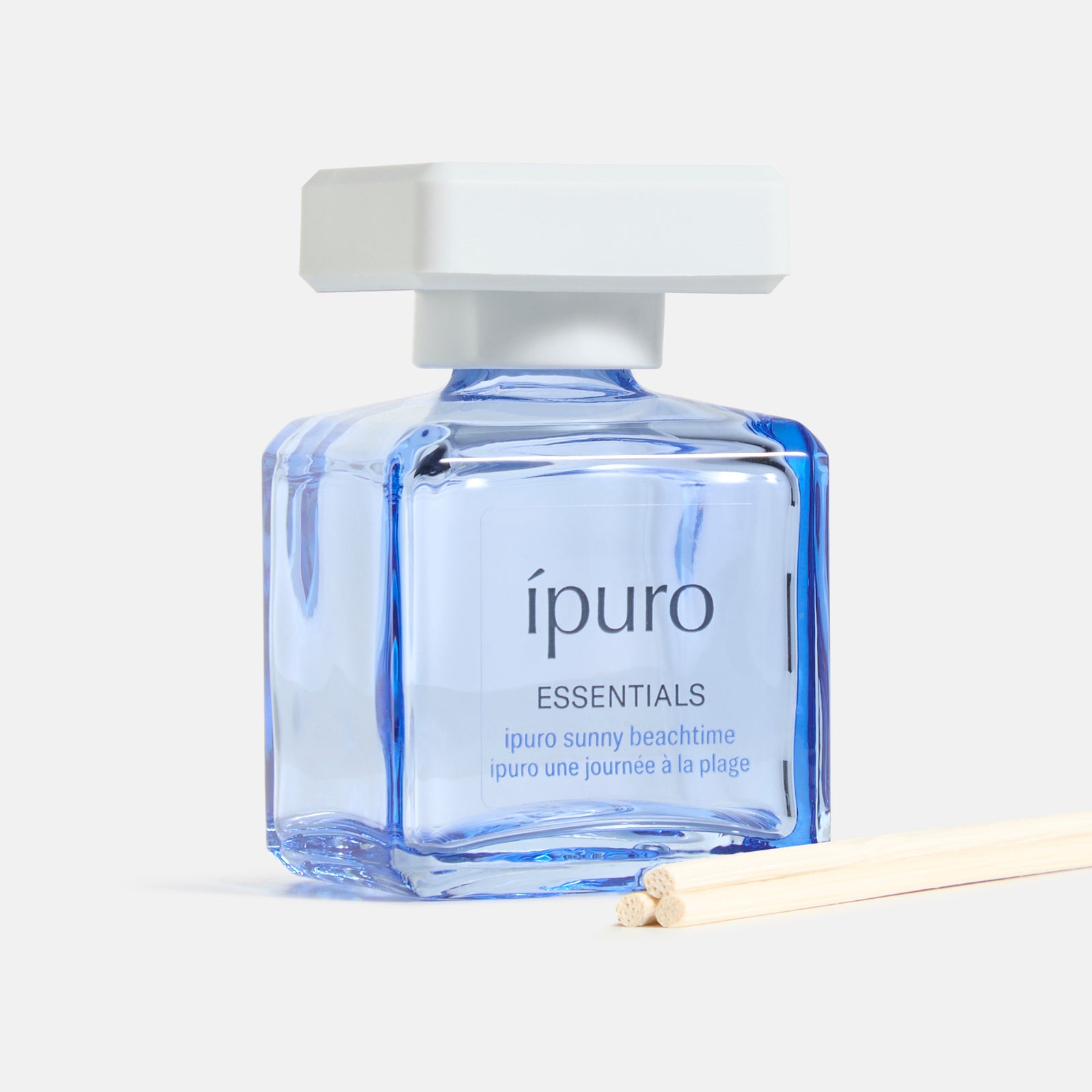 Eine quadratische Glasflasche von ipuro Raumduft sunny beachtime diffuser mit weißem Verschluss und hellblauer Flüssigkeit ist neben zwei Holzstäbchen auf hellem Hintergrund abgebildet.