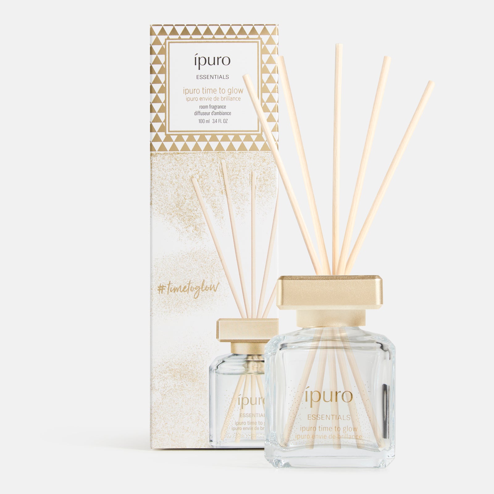Ein Glasflakon von ipuro time to glow Raumduft mit Goldkappe und Holzschilf wird vor der dazu passenden gold-weißen Schachtel gezeigt, die geometrische Muster, Produktdetails und Anklänge an Tonkabohne und Kaschmirholz aufweist.