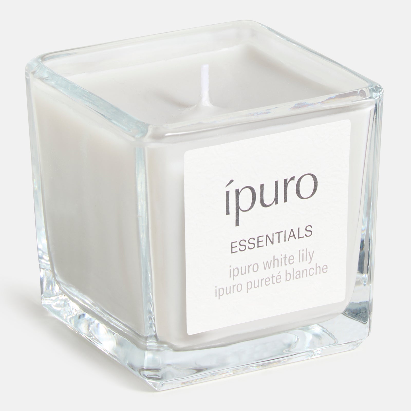 Die ipuro Duftkerze Weiße Lilie ist eine weiße Kerze in einem klaren, quadratischen Glasbehälter mit der Aufschrift "ipuro ESSENTIALS, ipuro Weiße Lilie, ipuro Pureté Blanche", die einen zarten Blumenduft verströmt.