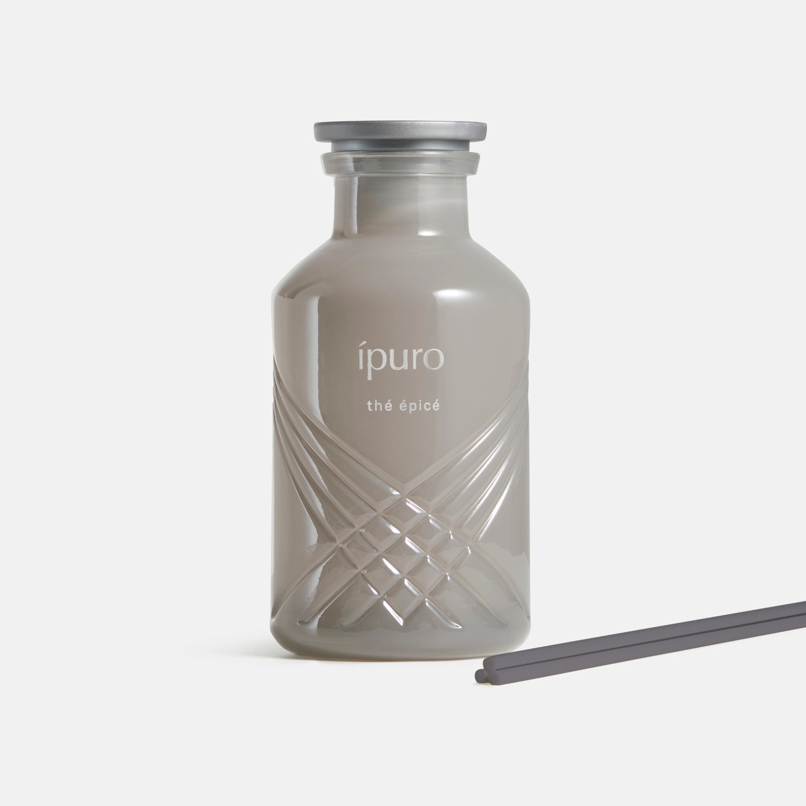 Der graue Glasflakon mit der Aufschrift "ipuro thé epicé" von ipuro hat einen silbernen Verschluss und eine kreuzförmige Textur. Jasmin-Noten duften die Luft, während zwei dunkle Diffusorstäbchen auf weißem Hintergrund daneben liegen.