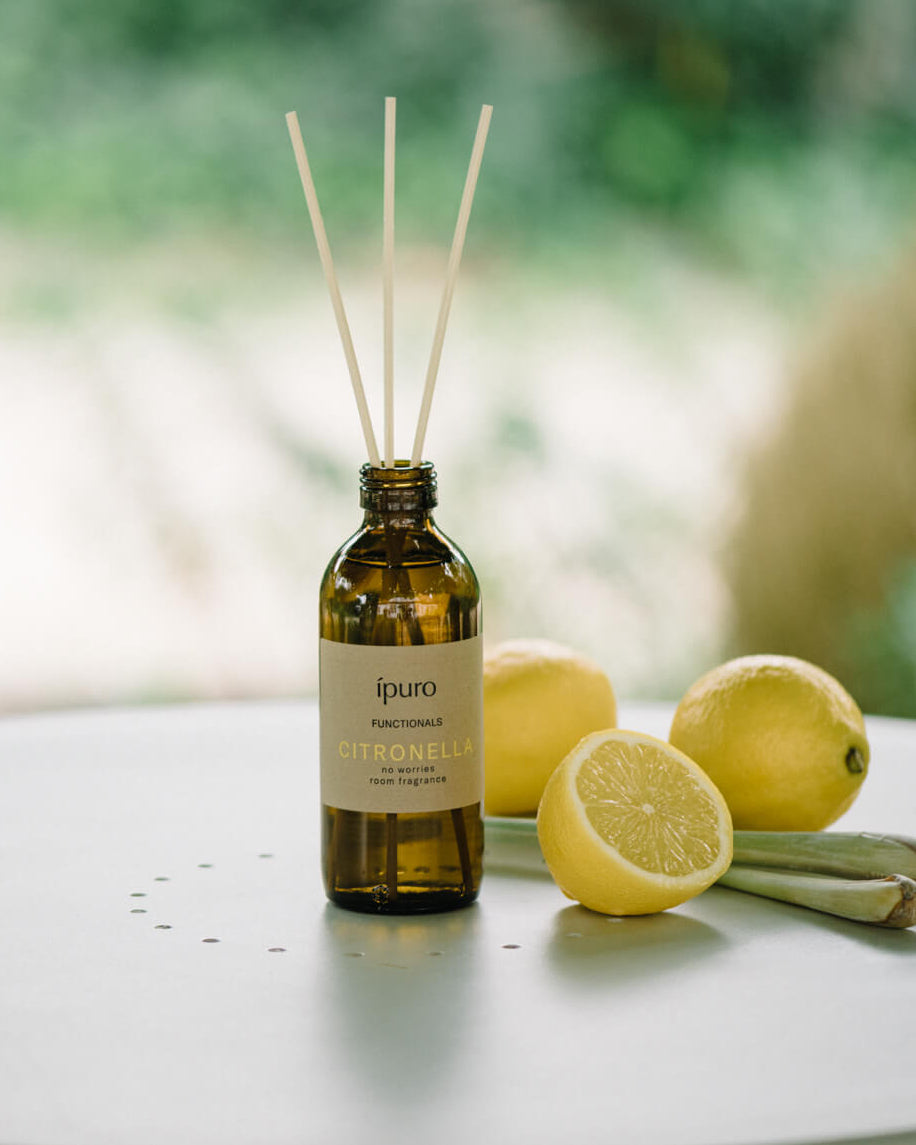 Eine braune Glasflasche mit der Aufschrift ipuro Citronella und Schilfrohr-Diffusorstäbchen steht auf einer weißen Fläche neben ganzen und halbierten Zitronen, mit einem verschwommenen grünen Hintergrund.