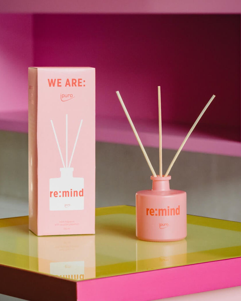 Ein rosafarbener Schilfrohrdiffusor mit der Aufschrift re:mind ipuro steht neben der dazugehörigen Box auf einer reflektierenden gelben und rosafarbenen Oberfläche mit einem leuchtend rosa Hintergrund. In dem Diffusor befinden sich drei Schilfrohre.