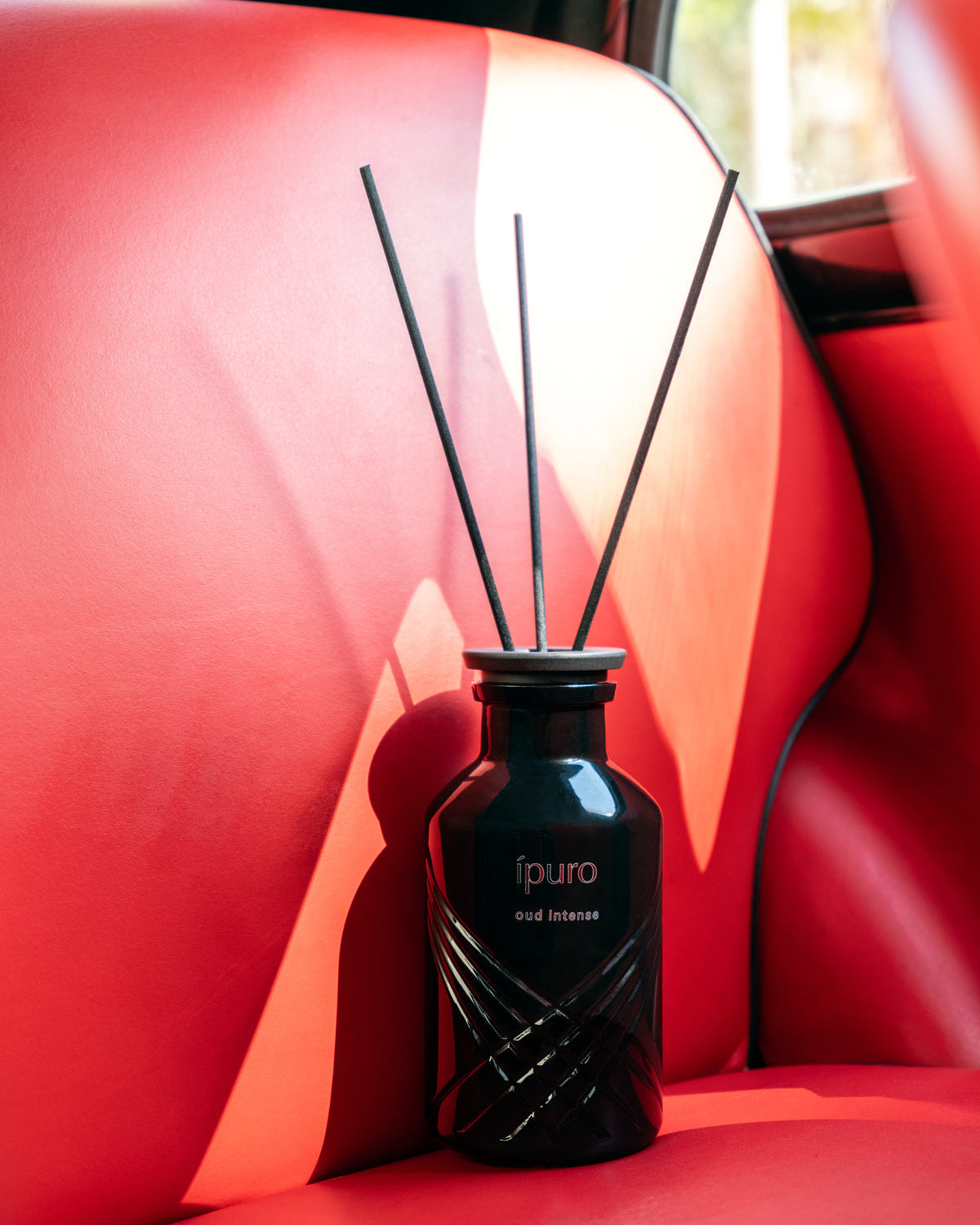 Ein schwarzer Ipuro Oud Intense Schilfrohrdiffusor mit drei Stäbchen auf einem leuchtend roten Ledersitz im Auto, während die Sonne aus dem Fenster scheint.