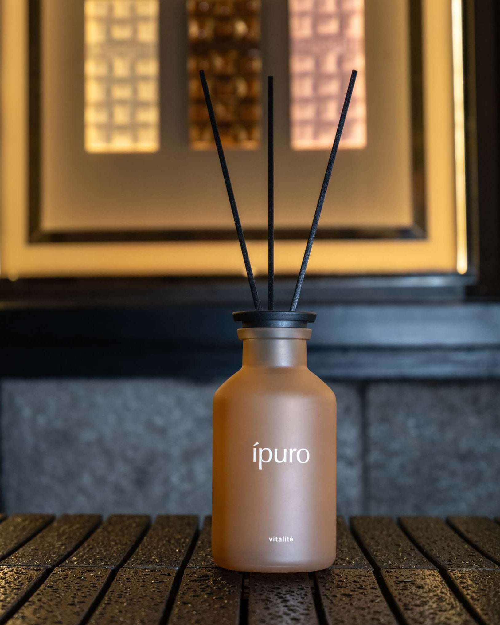 Eine braune Milchglasflasche mit der Aufschrift "ípuro vitalité" mit drei schwarzen Diffusorstäben, die auf einer strukturierten dunklen Oberfläche mit einem verschwommenen beleuchteten Hintergrund platziert sind.