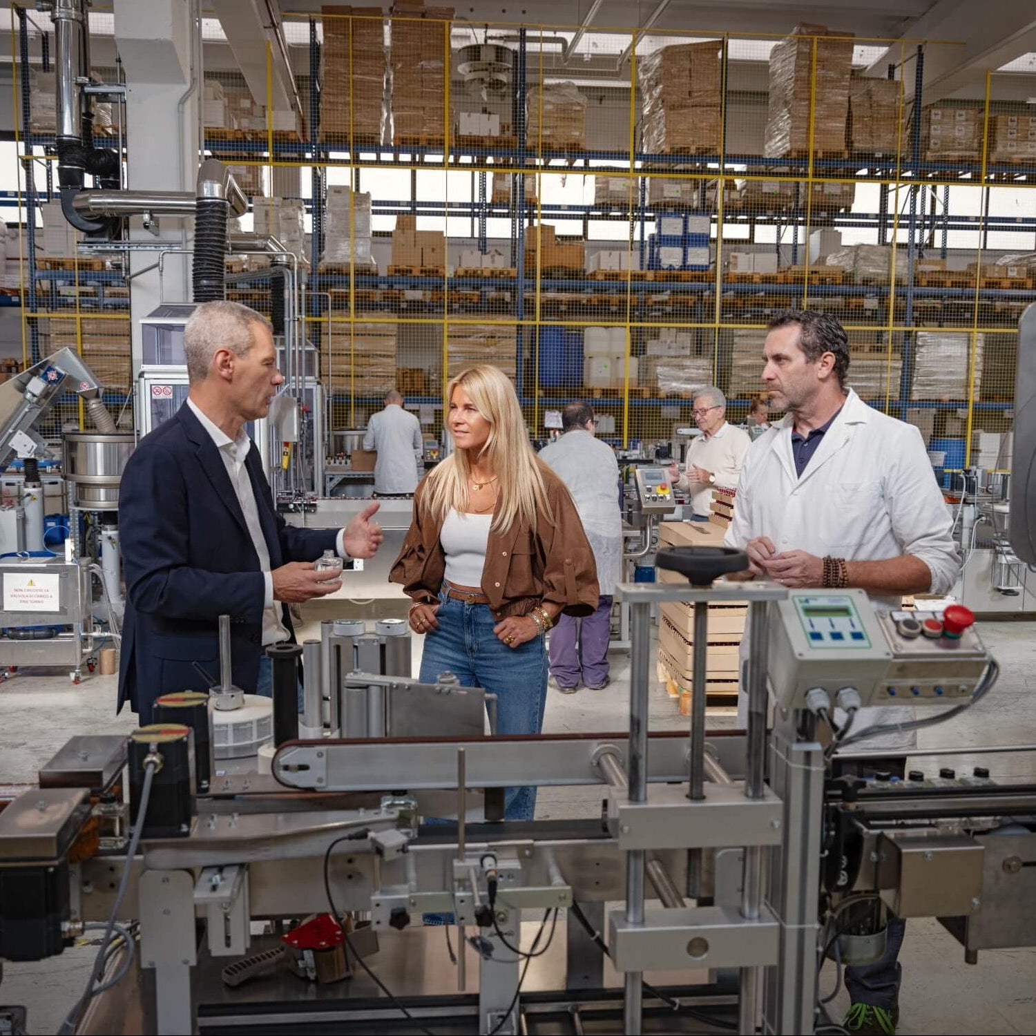 Drei Personen stehen und unterhalten sich in einer geschäftigen Fabrik mit Maschinen, Regalen mit Kisten und Arbeitern im Hintergrund. Die zwei Männer und die eine Frau scheinen etwas zu besprechen, das mit dem Produktionsprozess zu tun hat.