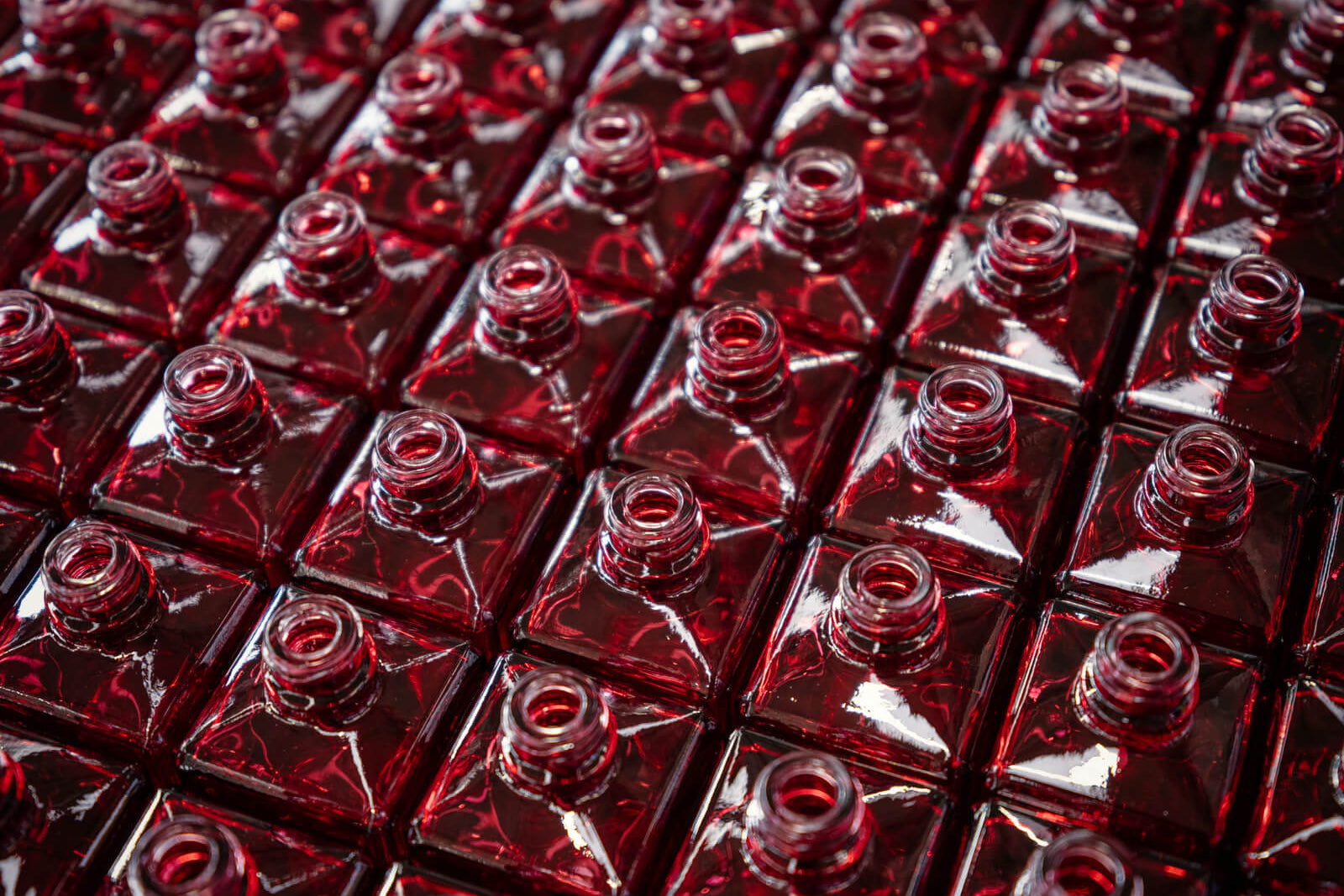 Reihen von quadratischen, offenen Glasflaschen, die mit roter Flüssigkeit gefüllt sind, sind von oben betrachtet eng aneinandergereiht und bilden ein geometrisches Muster mit Reflexionen und Glanzlichtern auf ihren Oberflächen.