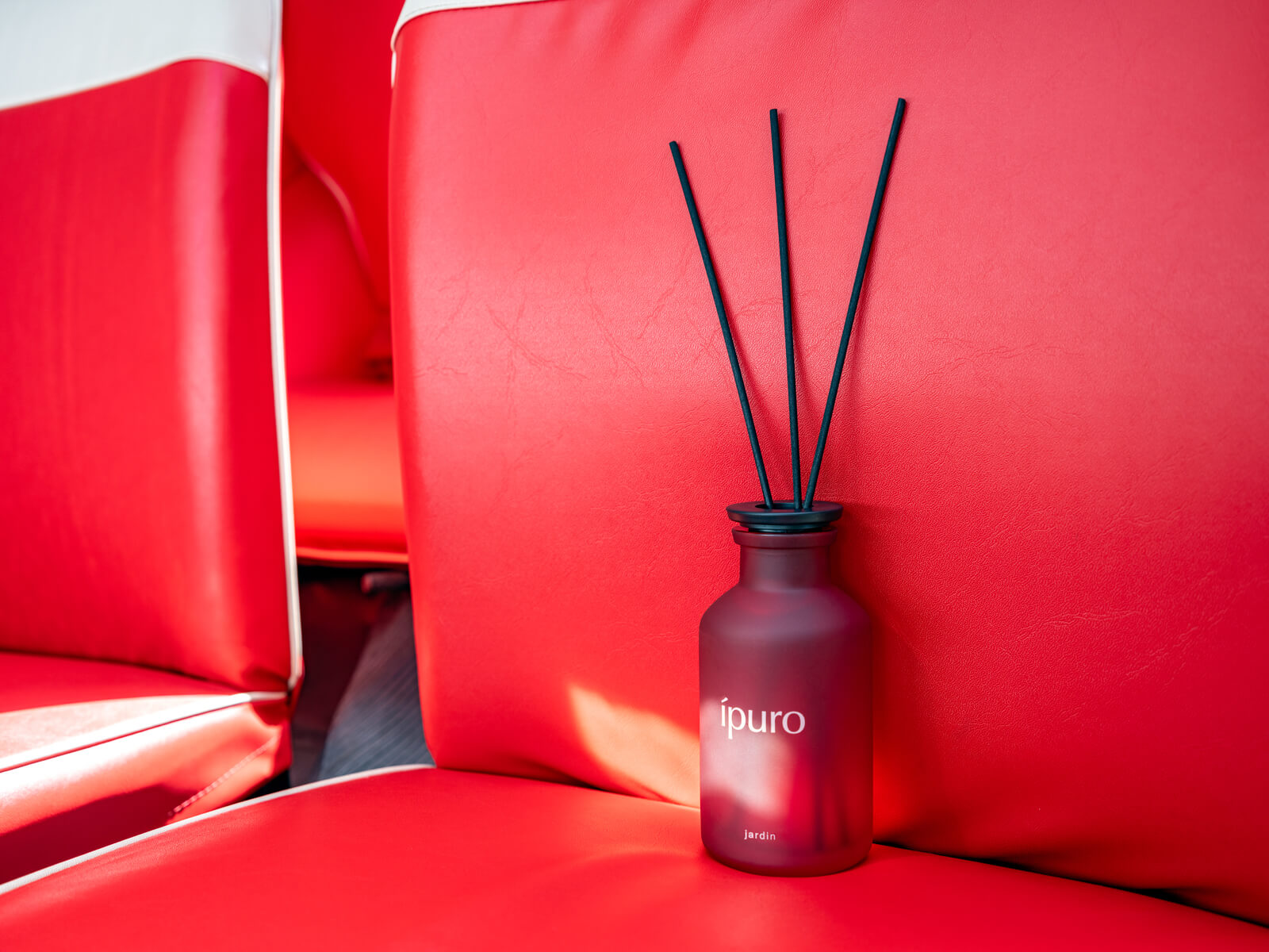 Ein roter Ipuro-Schilfrohrdiffusor mit schwarzen Stäben wird auf einen leuchtend roten und weißen Ledersitz gestellt und fängt das Sonnenlicht ein.