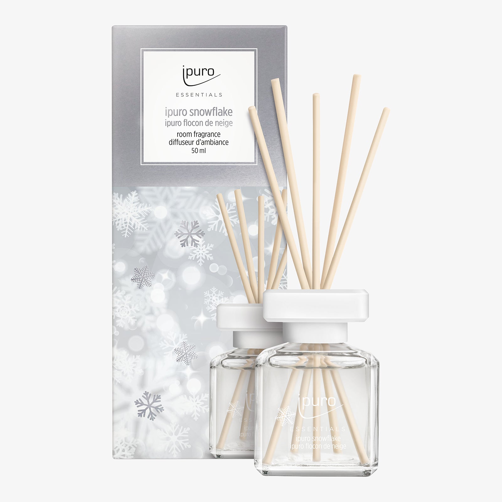 Zwei gläserne Diffusorflaschen mit weißen Kappen und Holzschilfrohr stehen vor einer silber-weißen ipuro ESSENTIALS Raumduft Snowflake Box, die mit Schneeflockenmotiven versehen ist und einen gemütlichen Winterduft verströmt.